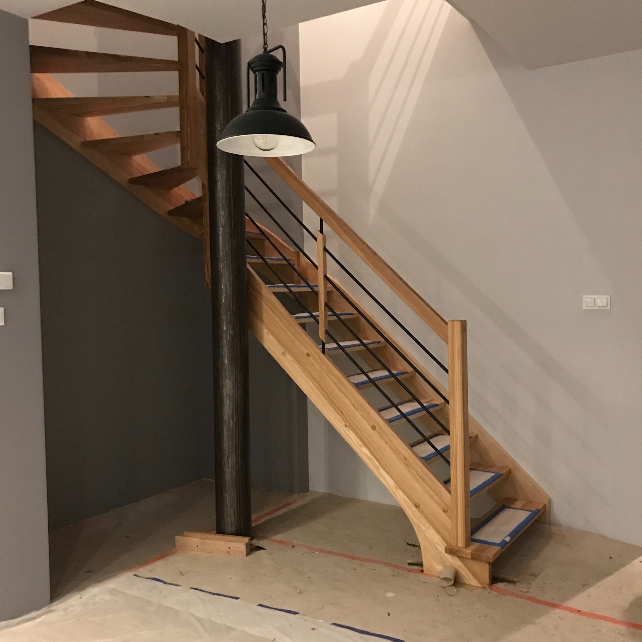 Surowe drewniane schody z czarnymi metalowymi balustradami, wsparte na ciemnym, okrągłym słupie, z prowizorycznymi zabezpieczeniami na stopniach, pod wiszącą lampą w industrialnym stylu, w trakcie...