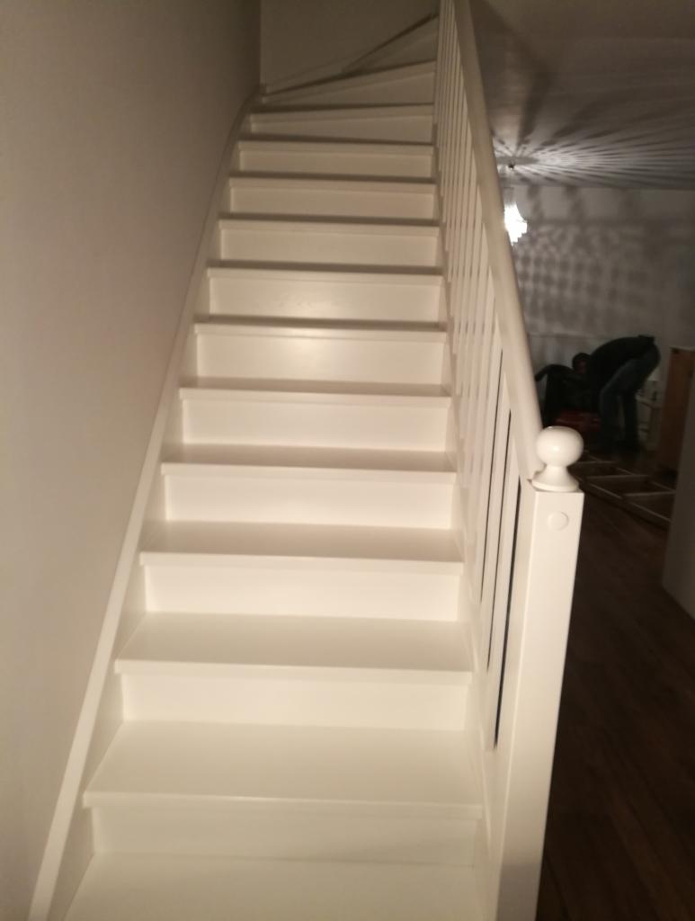 Białe schody z drewnianą balustradą w jasnym wnętrzu, widok od dołu, w tle osoba przy pracy.