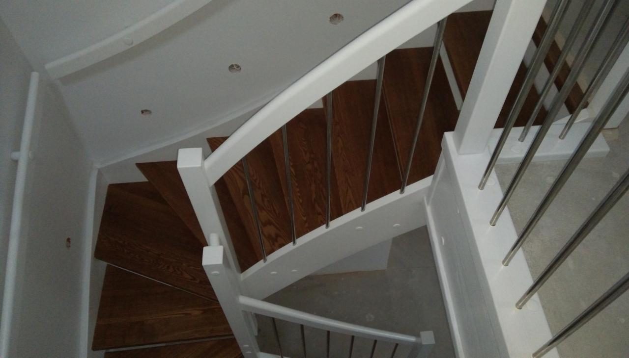 Widok z góry na drewniane schody spiralne z białymi balustradami i metalowymi prętami, oświetlenie sufitowe punktowe.