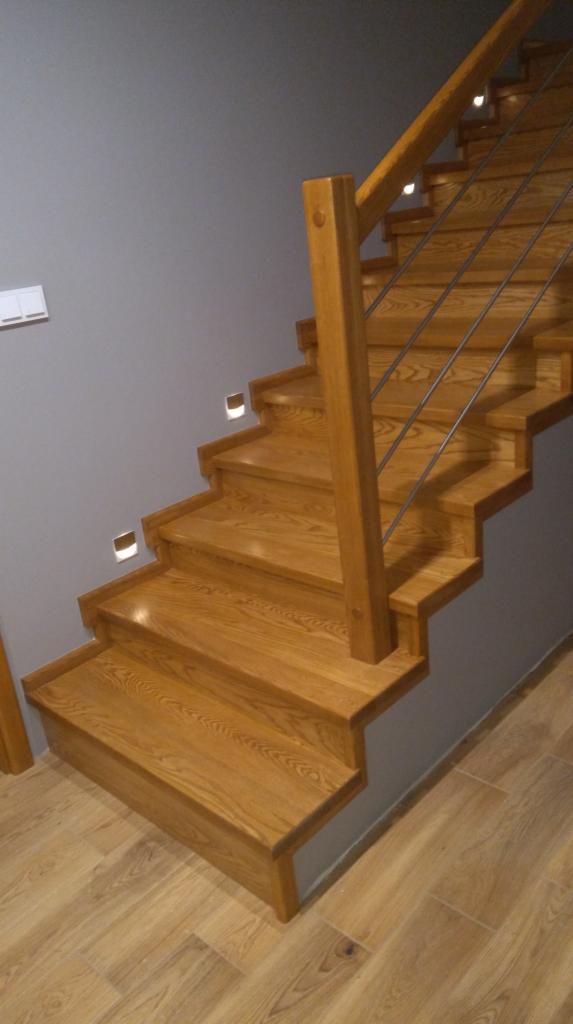 Dębowe schody z metalowymi prętami balustrady i oświetleniem punktowym w ścianie, widok z dołu na pierwsze stopnie.