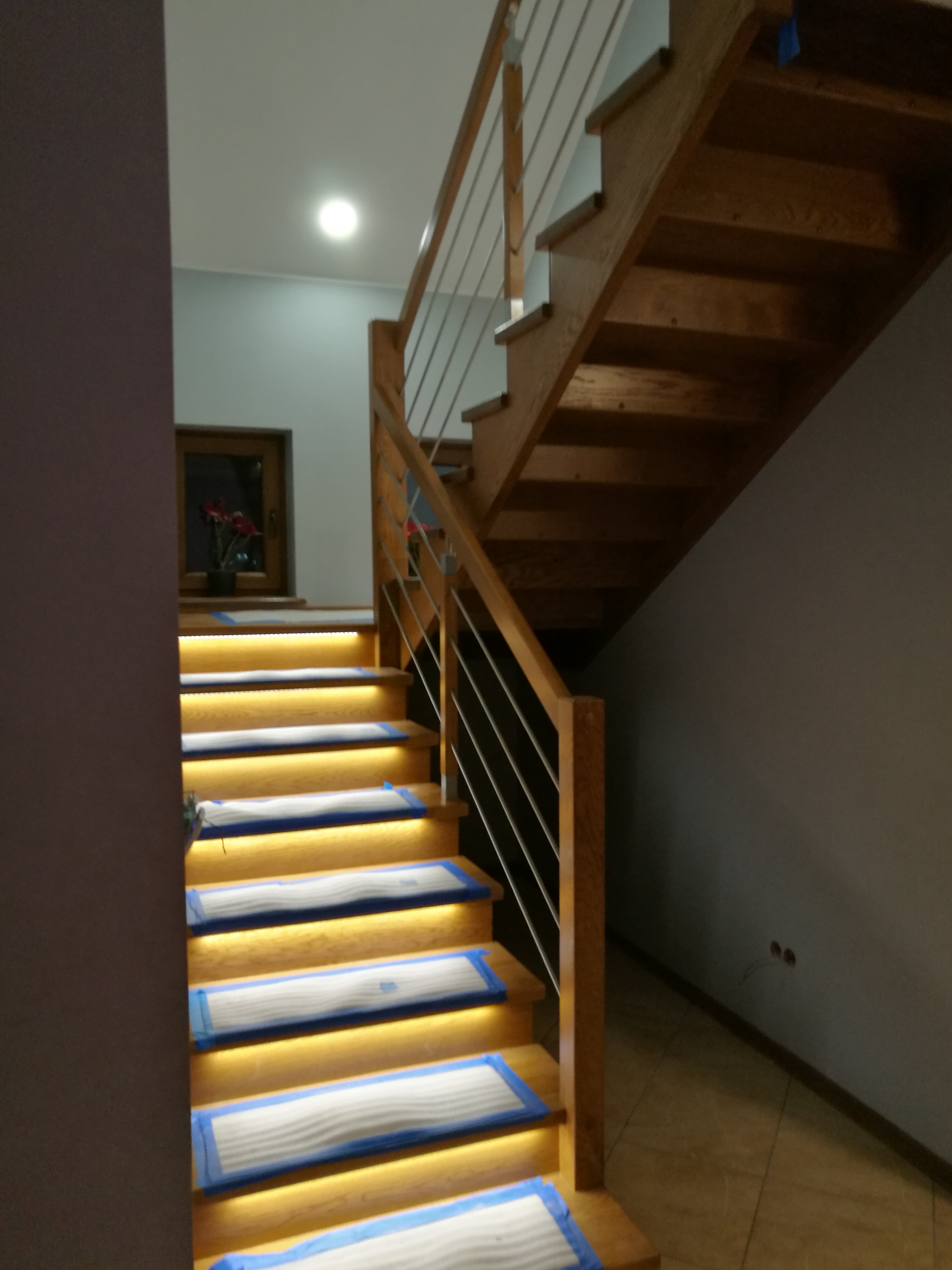 Nowoczesne schody z drewna z podświetleniem LED na stopniach, zabezpieczone folią malarską, metalowa balustrada, widok z dołu.