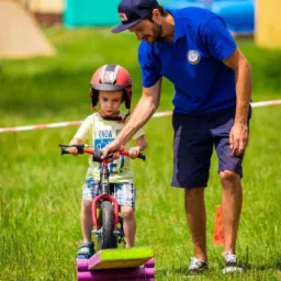 Animator w niebieskiej koszulce uczy małego chłopca w kasku i koszulce z napisem 'I KNOW Y' jazdy na rowerku biegowym na trawie, w tle dmuchane zamki i toalety przenośne.