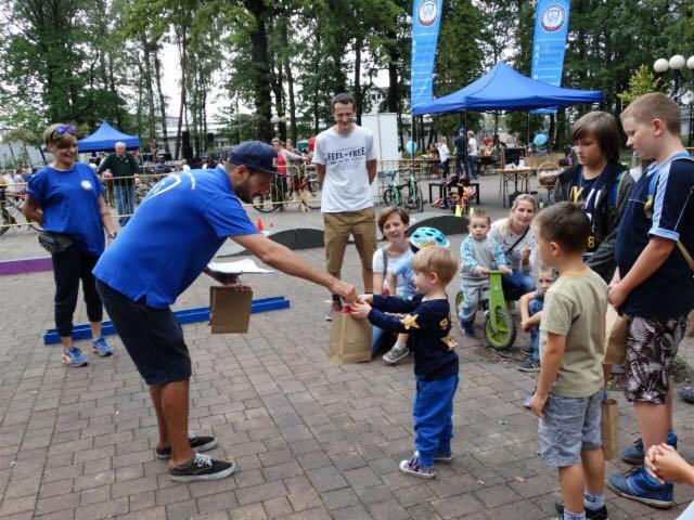 Wręczenie nagród w papierowych torbach małemu chłopcu w niebieskim dresie podczas plenerowej imprezy dla dzieci, w tle widoczni rodzice i inne dzieci.
