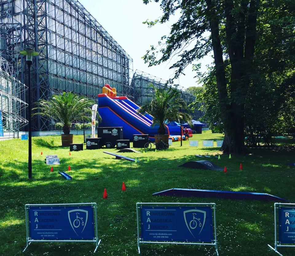 Zewnętrzna strefa zabaw dla dzieci z dmuchaną zjeżdżalnią, rampami rowerowymi i palmami w donicach na trawie, przed nowoczesnym budynkiem ze szklaną fasadą, w tle widoczne namioty.
