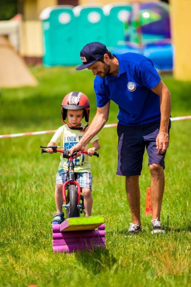 Animator w niebieskiej koszulce uczy małego chłopca w kasku i koszulce z napisem 'I KNOW Y' jazdy na rowerku biegowym na trawie, w tle dmuchane zamki i toalety przenośne.