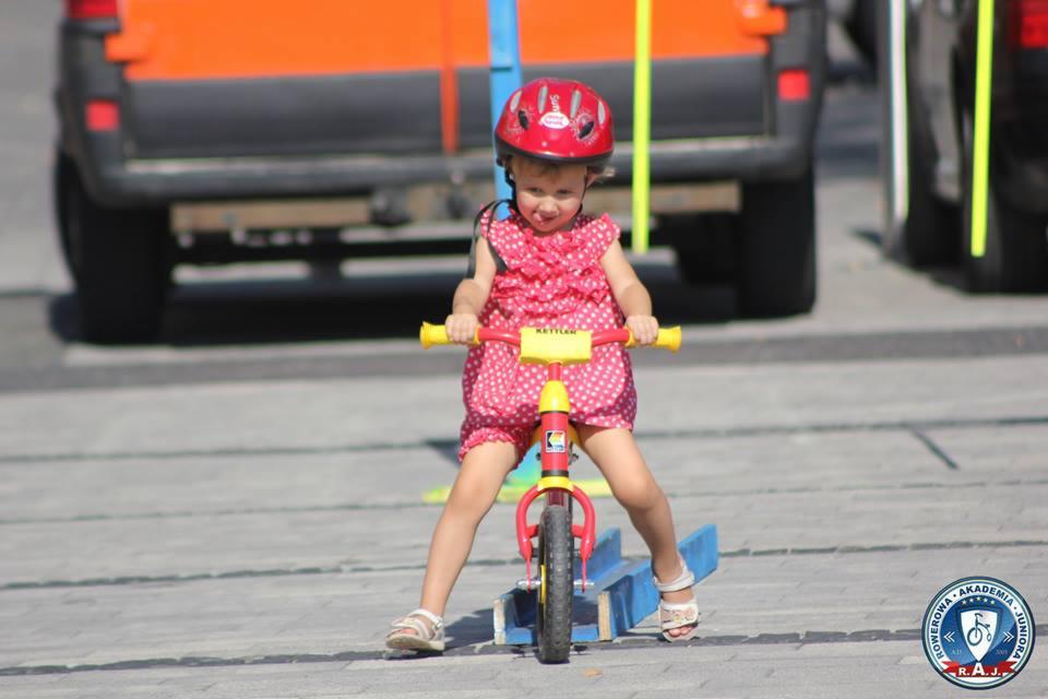 Dziewczynka w czerwonym kasku i różowej sukience w białe kropki uczy się jeździć na czerwono-żółtym rowerku biegowym Kettler na tle pomarańczowego busa.