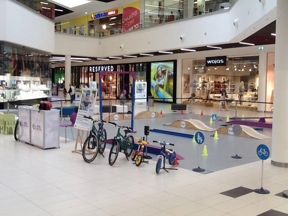 Tymczasowy tor rowerowy dla dzieci w centrum handlowym, z małymi rampami, znakami drogowymi i rowerami dla dzieci w różnych rozmiarach.