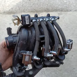 Czarny kolektor dolotowy z zamontowaną instalacją gazową LPG, widoczne wtryskiwacze gazu, przewody i opaski zaciskowe, trzymany w dłoni na tle betonowej nawierzchni.