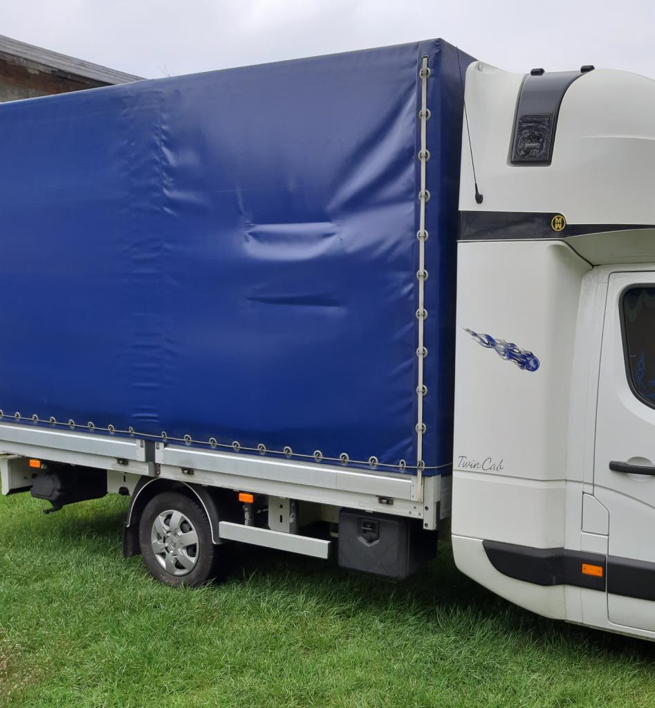 Biały samochód dostawczy z niebieską plandeką na trawie, widoczny napis 'Twin Cab' i grafika przypominająca kometę na kabinie.