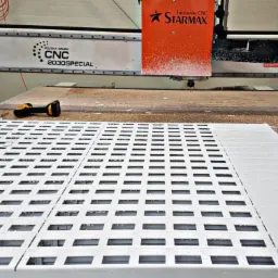 Proces obróbki CNC białej, ażurowej konstrukcji, prawdopodobnie elementu drzwi lub mebla, widoczny frez i opiłki, w tle maszyna CNC Starmx.