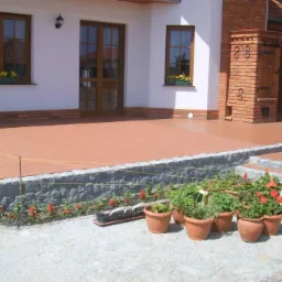 Wykończony taras z czerwonej kostki brukowej przy białym domu z ceglanymi elementami, widoczne donice z kwiatami i mały ogródek na pierwszym planie.