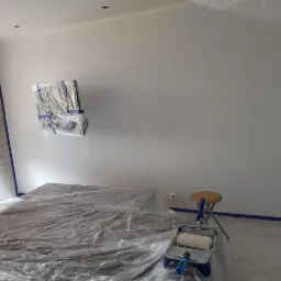 Pokój przygotowany do malowania: meble i podłoga zabezpieczone folią, ściany oklejone taśmą malarską, wałek i kuweta malarska obok krzesła.