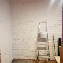 Ściana wykończona białymi płytkami imitującymi cegłę, z metalową drabiną opartą o nią, widoczne ślady prac wykończeniowych na podłodze.