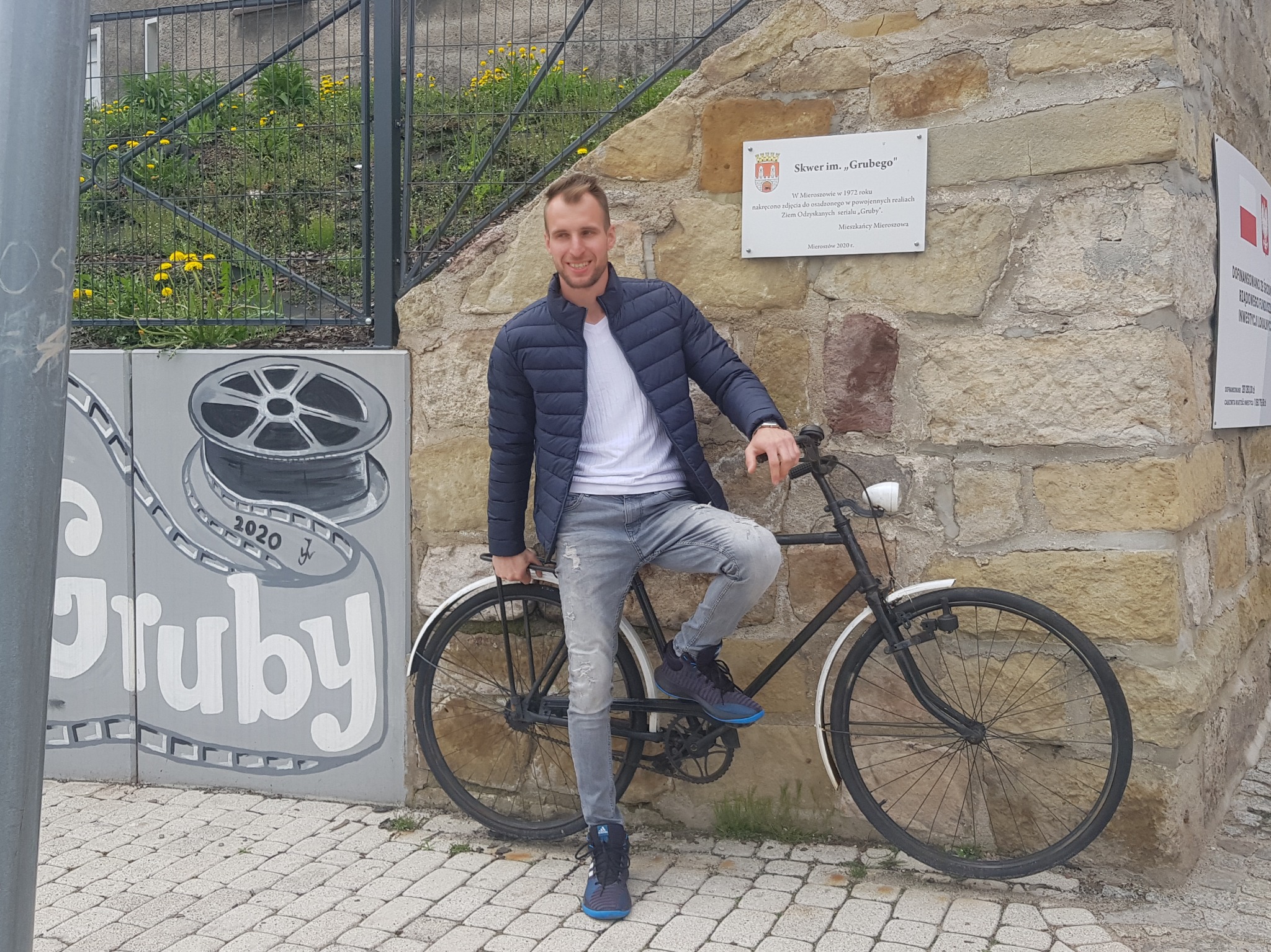 Uśmiechnięty mężczyzna w kurtce pozuje z rowerem przy kamiennej ścianie z tablicą informacyjną i muralem nawiązującym do filmu.