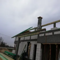 Wznoszenie ścian domu murowanego z betonu komórkowego, widoczne kominy w trakcie budowy, drewniane elementy konstrukcyjne dachu i podparcia, deski składowane obok budynku.