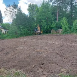 Przygotowany teren pod trawnik z widocznym traktorem kopiącym w tle, obok drewniana furtka i zieleń drzew.