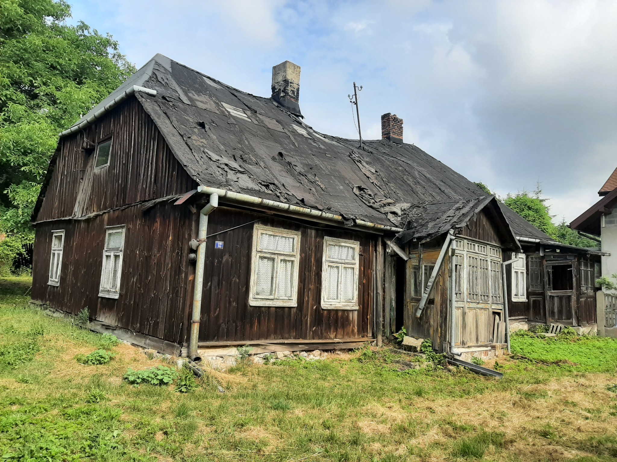Stary, drewniany dom z mocno zniszczonym dachem pokrytym papą, widoczne dwa kominy, zarośnięty ogród, elewacja z ciemnego drewna.