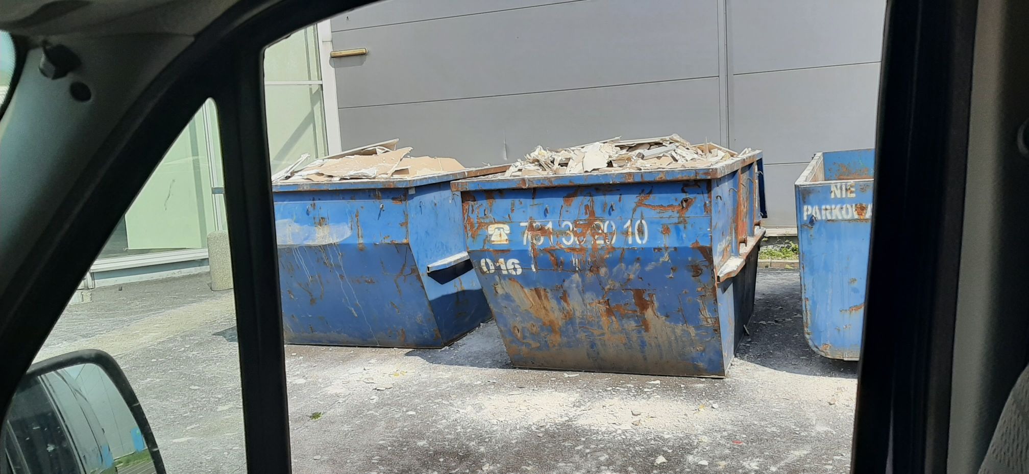 Trzy niebieskie, zużyte kontenery na gruz wypełnione płytami gipsowymi i innymi odpadami budowlanymi, widziane z wnętrza pojazdu przez otwarte okno.