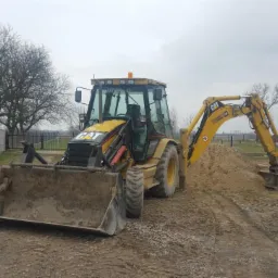 Żółta koparko-ładowarka CAT na placu budowy z pryzmą ziemi, widoczne ślady opon i betonowe ogrodzenie w tle.