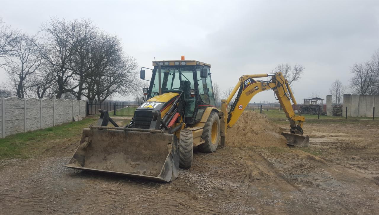 Żółta koparko-ładowarka CAT na placu budowy z pryzmą ziemi, widoczne ślady opon i betonowe ogrodzenie w tle.