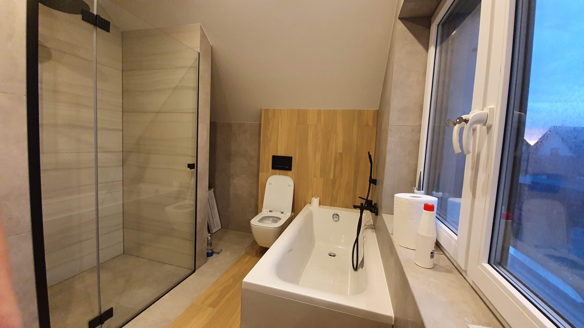 Nowoczesna łazienka na poddaszu z kabiną prysznicową typu walk-in z czarnymi profilami, białą wanną, toaletą i drewnianą okładziną ścienną.