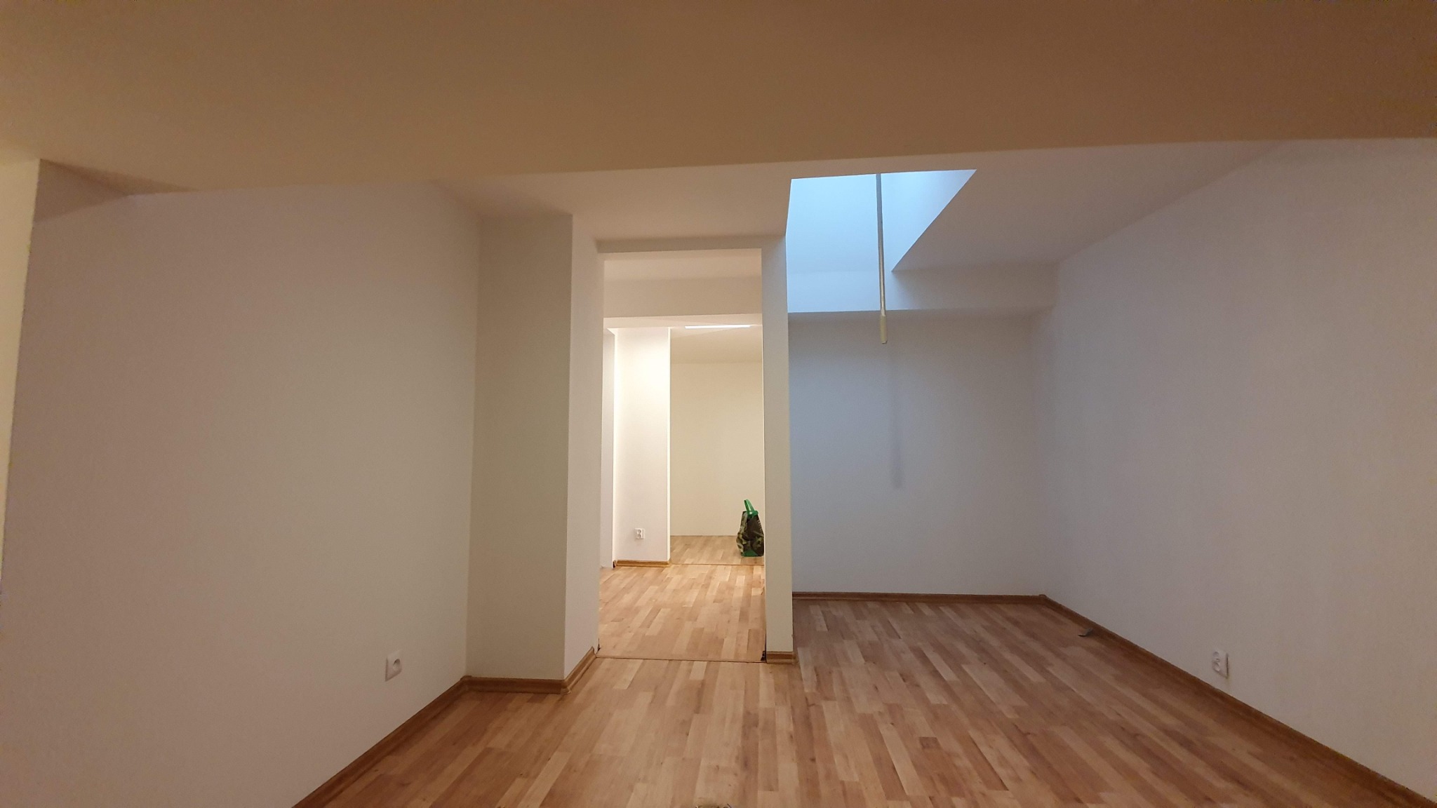 Puste, odświeżone wnętrze z drewnianą podłogą i świeżo pomalowanymi na biało ścianami, widok na kolejne pomieszczenie przez otwarty prześwit, z torbą stojącą w tle.