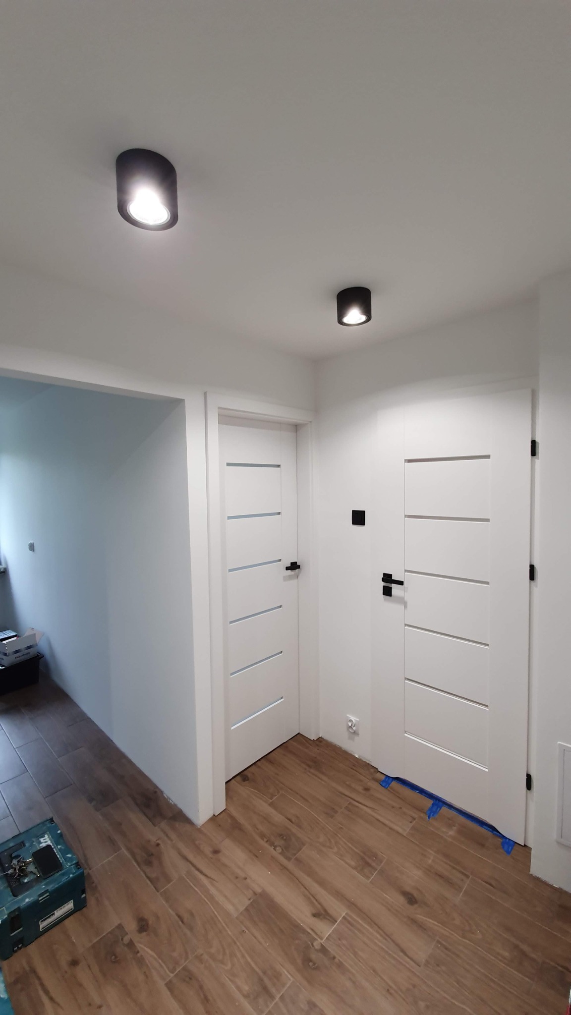 Korytarz po remoncie z drewnopodobną podłogą, białymi drzwiami z poziomymi frezami i czarnymi klamkami, oświetlony dwoma czarnymi lampami sufitowymi, z widoczną skrzynką narzędziową w rogu.