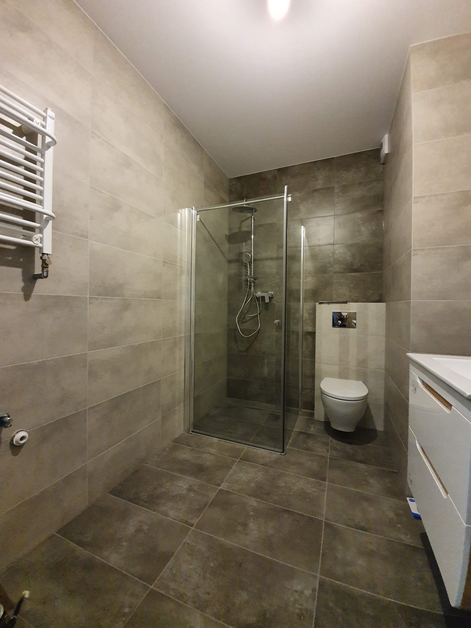 Nowoczesna łazienka z kabiną prysznicową, grzejnikiem drabinkowym i umywalką z szafką w kolorze białym, wykończona płytkami w odcieniach szarości.