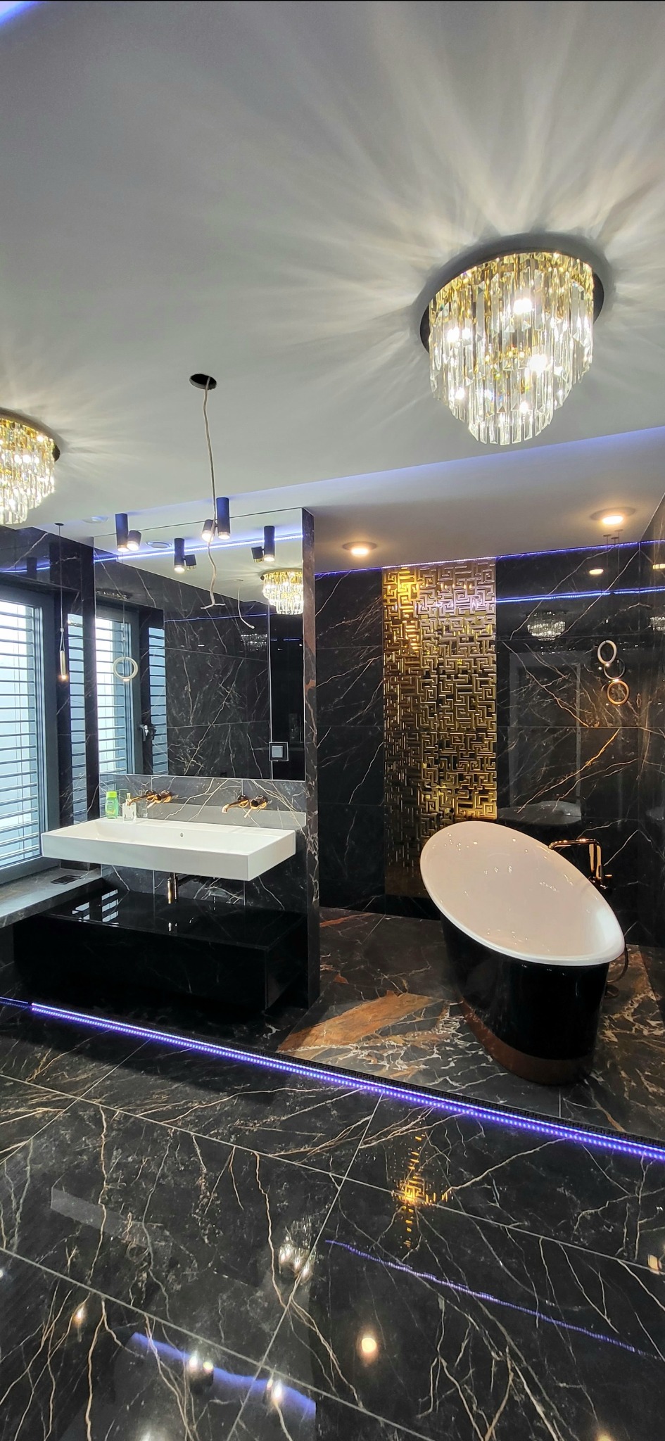 Luksusowa łazienka z czarnymi marmurowymi płytkami, wolnostojącą białą wanną na czarnej podstawie, podwieszaną białą umywalką z złotą armaturą, złotą dekoracyjną ścianką działową i kryształowymi...