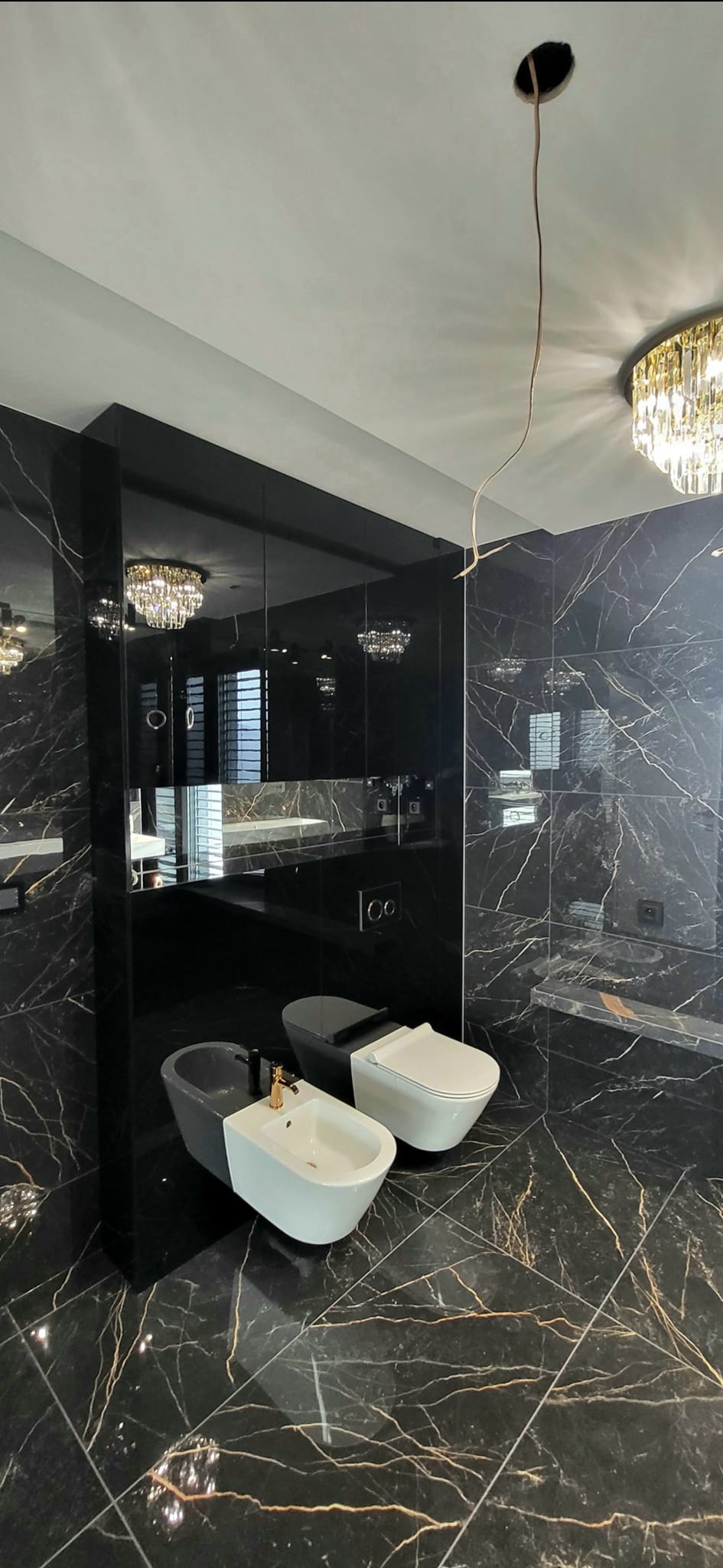 Luksusowa łazienka z czarnymi marmurowymi płytkami, wiszącą toaletą i bidetem w kolorze szaro-białym, lustrzaną zabudową i złotym żyrandolem, widoczne kable od oświetlenia sufitowego.
