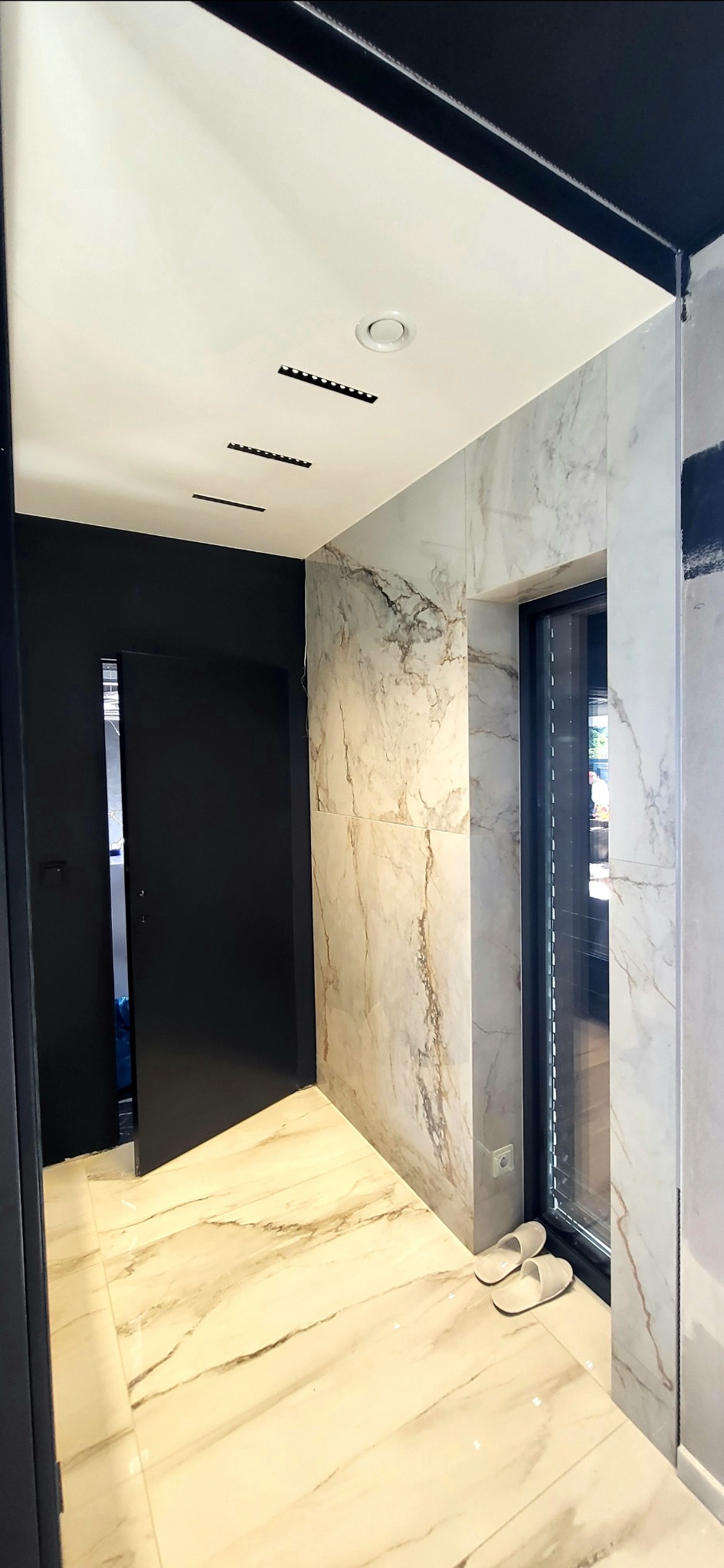 Nowoczesny korytarz z marmurowymi płytkami na podłodze i ścianach, czarne drzwi i oświetlenie punktowe w suficie, para białych kapci przy oknie.