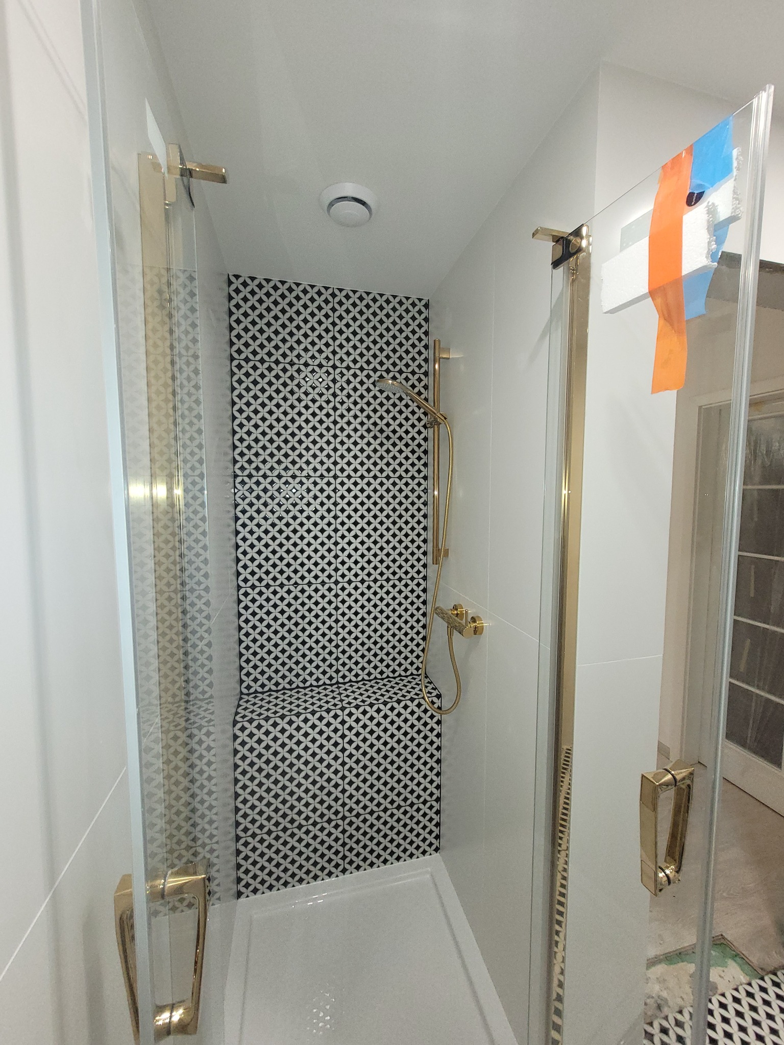 Nowoczesna kabina prysznicowa z geometrycznym wzorem płytek w odcieniach czerni i bieli, złote okucia, częściowo zabezpieczona folią malarską.