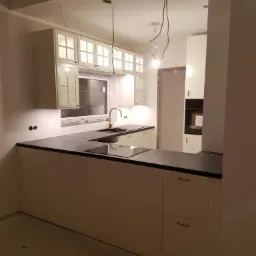 Wnętrze kuchni w trakcie wykańczania: białe szafki z przeszklonymi frontami, czarny blat i złota bateria, widoczne kable oświetleniowe