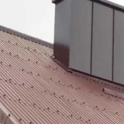 Ciemnobrązowy komin wentylacyjny na dachu z blachy trapezowej w kolorze ceglastym, widok z dołu pod kątem, szare niebo w tle.