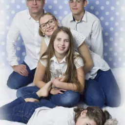 Portret rodzinny: rodzice i trójka dzieci w jasnych ubraniach na tle w niebieskie kropki, najmłodsza córka leży z pluszowym misiem.