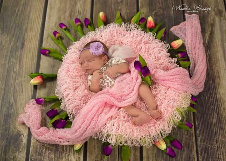 Śpiąca noworodka w różowym gniazdku z tulipanami na drewnianym tle, fioletowa opaska z kwiatkiem na główce.