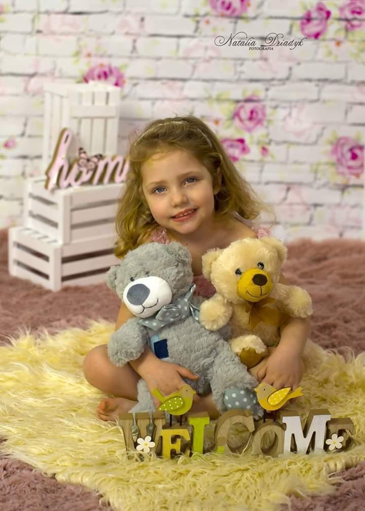 Uśmiechnięta dziewczynka z blond włosami siedzi na puszystym dywanie trzymając dwa pluszowe misie na tle ściany w kwiaty, przed nią napis 'Welcome' z drewnianymi ptaszkami.