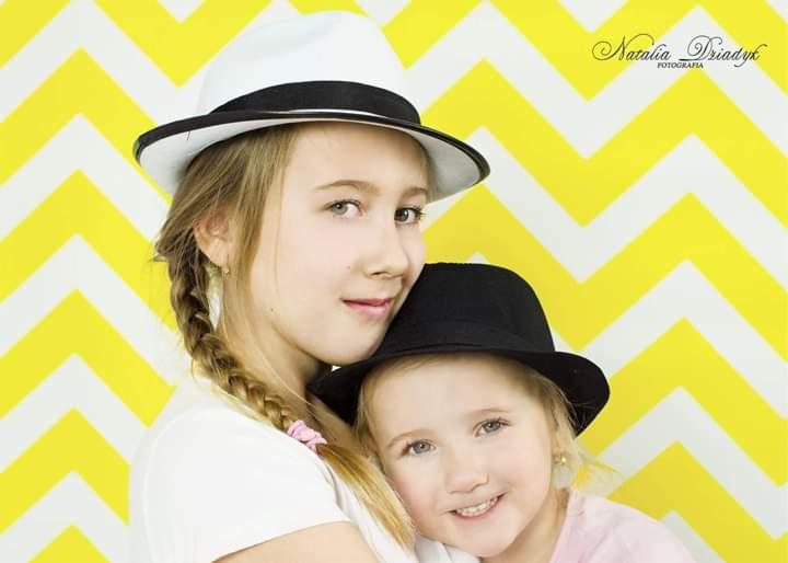 Portret dwóch uśmiechniętych dziewczynek w kapeluszach na tle żółto-białego wzoru chevron, starsza z warkoczem i białym kapeluszu, młodsza w czarnym kapeluszu.