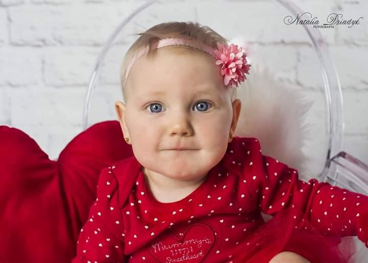 Portret niemowlęcia z niebieskimi oczami, w czerwonym ubranku w białe serduszka, z różową opaską i kwiatem na głowie, siedzącego w przezroczystym krześle na tle białej ceglanej ściany.