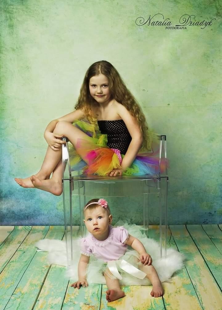 Portret dwóch dziewczynek w studio: starsza siedzi na przezroczystym krześle w czarnej sukience w kropki i tęczowej spódniczce tutu, młodsza siedzi na podłodze w różowej bluzce i białej spódniczce...