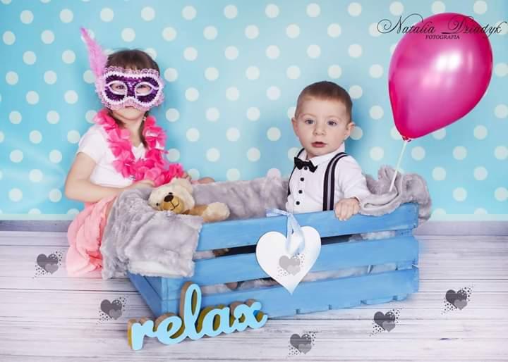 Dzieci w niebieskiej skrzynce na tle błękitnej ściany w kropki: dziewczynka w masce karnawałowej i różowym boa, chłopiec w koszuli z muchą i szelkami, różowy balon, pluszowy miś i napis 'relax'.