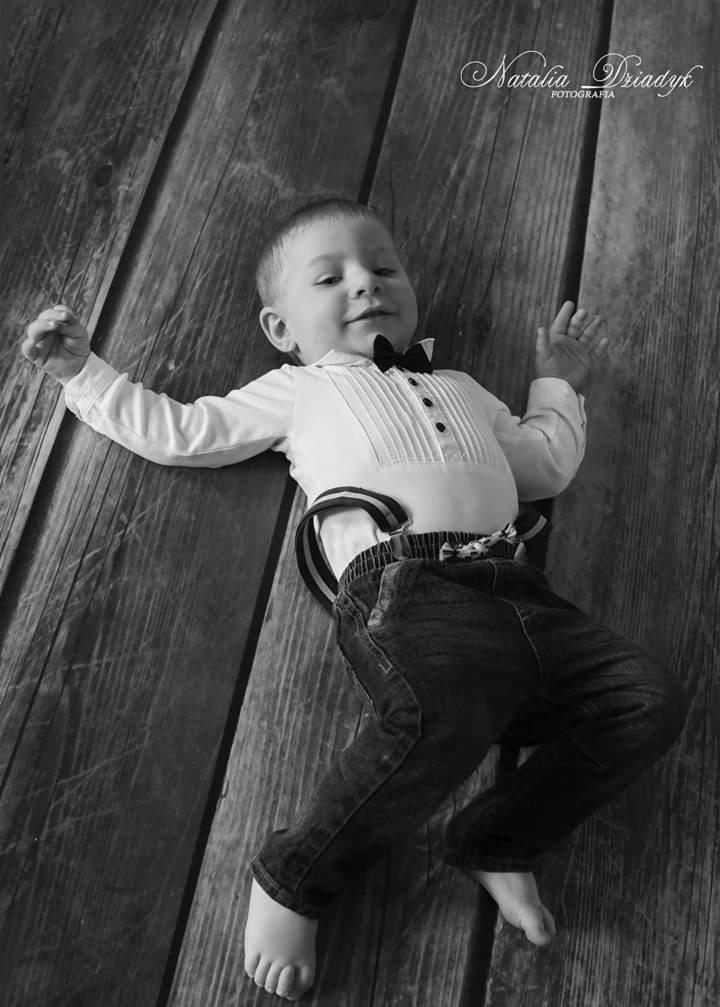 Czarno-białe zdjęcie małego chłopca w koszuli z muchą i jeansach z szelkami, leżącego na drewnianej podłodze, z logo fotografa w prawym górnym rogu.