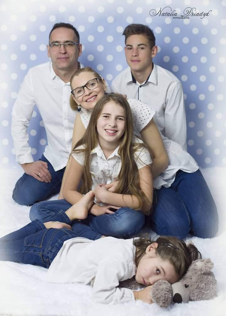 Portret rodzinny: rodzice i trójka dzieci w jasnych ubraniach na tle w niebieskie kropki, najmłodsza córka leży z pluszowym misiem.