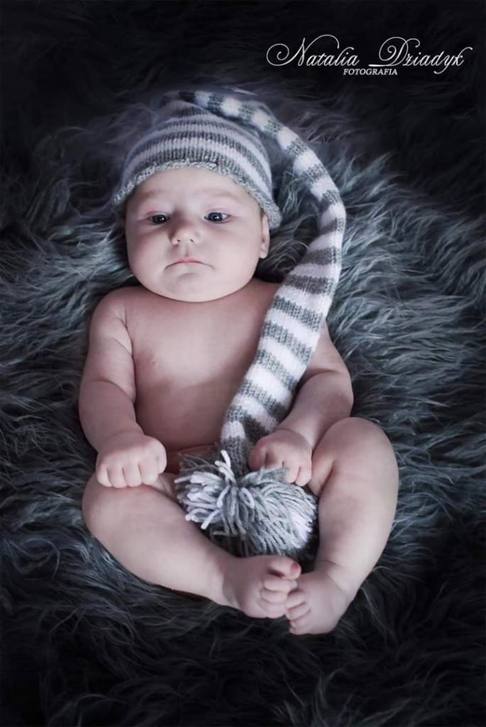 Noworodek w paskiwanym czepku z pomponem, leżący na szarym futrzanym tle, z delikatnym oświetleniem.