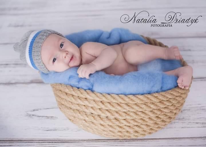 Uśmiechnięty noworodek w dzierganej czapce, leżący w koszyku wyłożonym błękitnym futerkiem, na tle białych desek.
