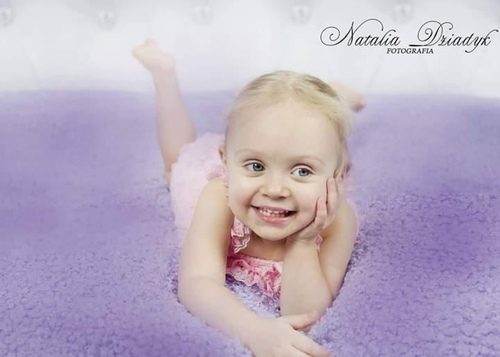 Uśmiechnięta blond dziewczynka w różowej sukience leży na fioletowym puszystym kocu, opierając głowę na dłoni. W tle białe tło i logo fotografa.