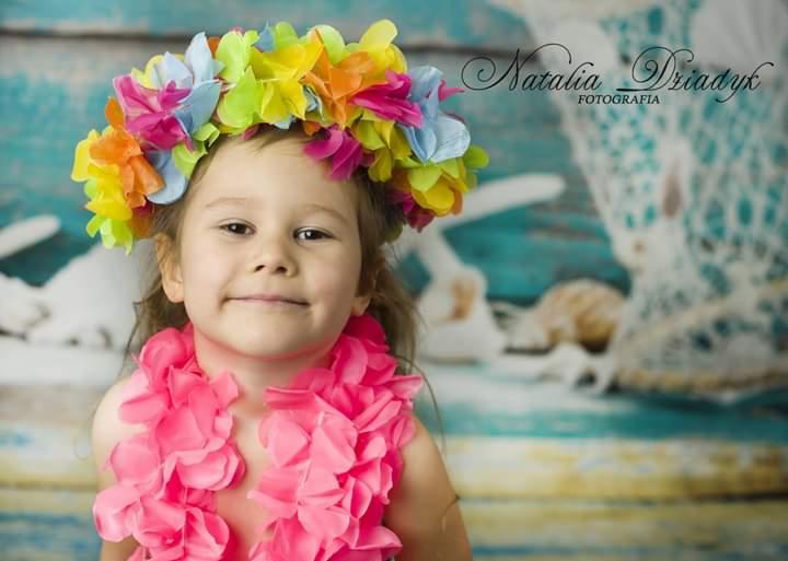 Portret uśmiechniętej dziewczynki z wiankiem z kolorowych kwiatów i różową hawajską girlandą na szyi, na tle stylizowanej, błękitnej ściany z elementami morskimi.