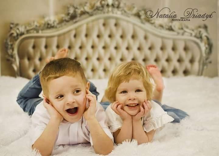 Uśmiechnięty chłopiec i dziewczynka leżący na puszystym białym kocu, podpierający twarze dłońmi, w tle ozdobny zagłówek łóżka i logo fotografa.