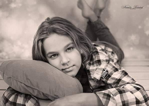 Portret czarno-biały nastolatki leżącej na poduszce w koszuli w kratę, z nogami uniesionymi do góry, na tle abstrakcyjnym.