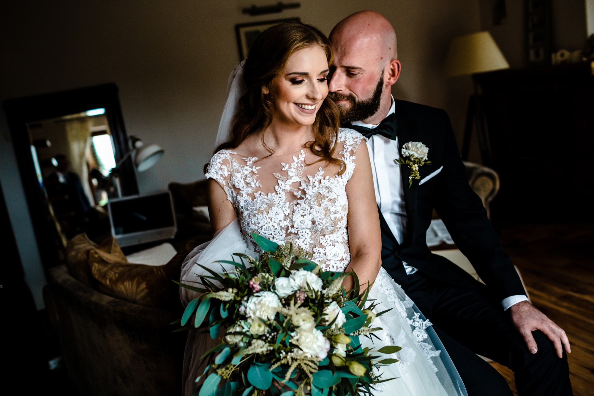 Uśmiechnięta panna młoda w koronkowej sukni trzyma bukiet ślubny, obok pan młody w czarnym smokingu z muszką.